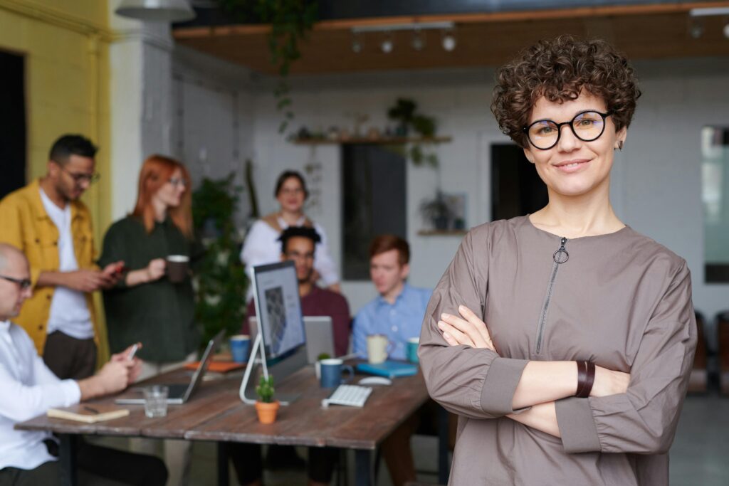 Smiling businesswoman with curly hair stands confidently in a modern office space with colleagues.